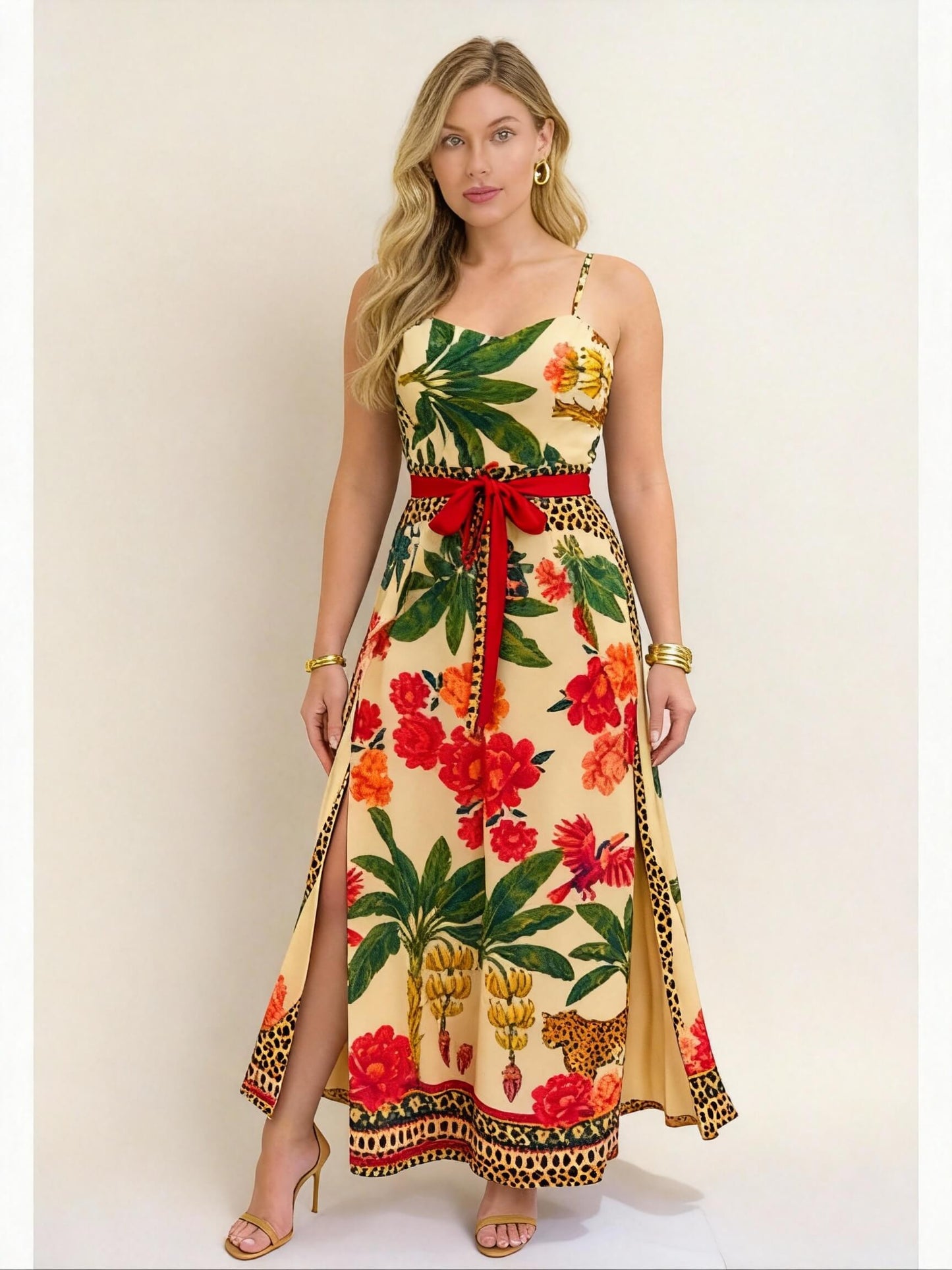 Modelo vestindo vestido longo de verão com estampa tropical vibrante em fundo amarelo, com folhas verdes, flores vermelhas, bananas e detalhes de oncinha. Cintura marcada com laço vermelho e fenda lateral, ideal para um look elegante e ousado em dias quentes.