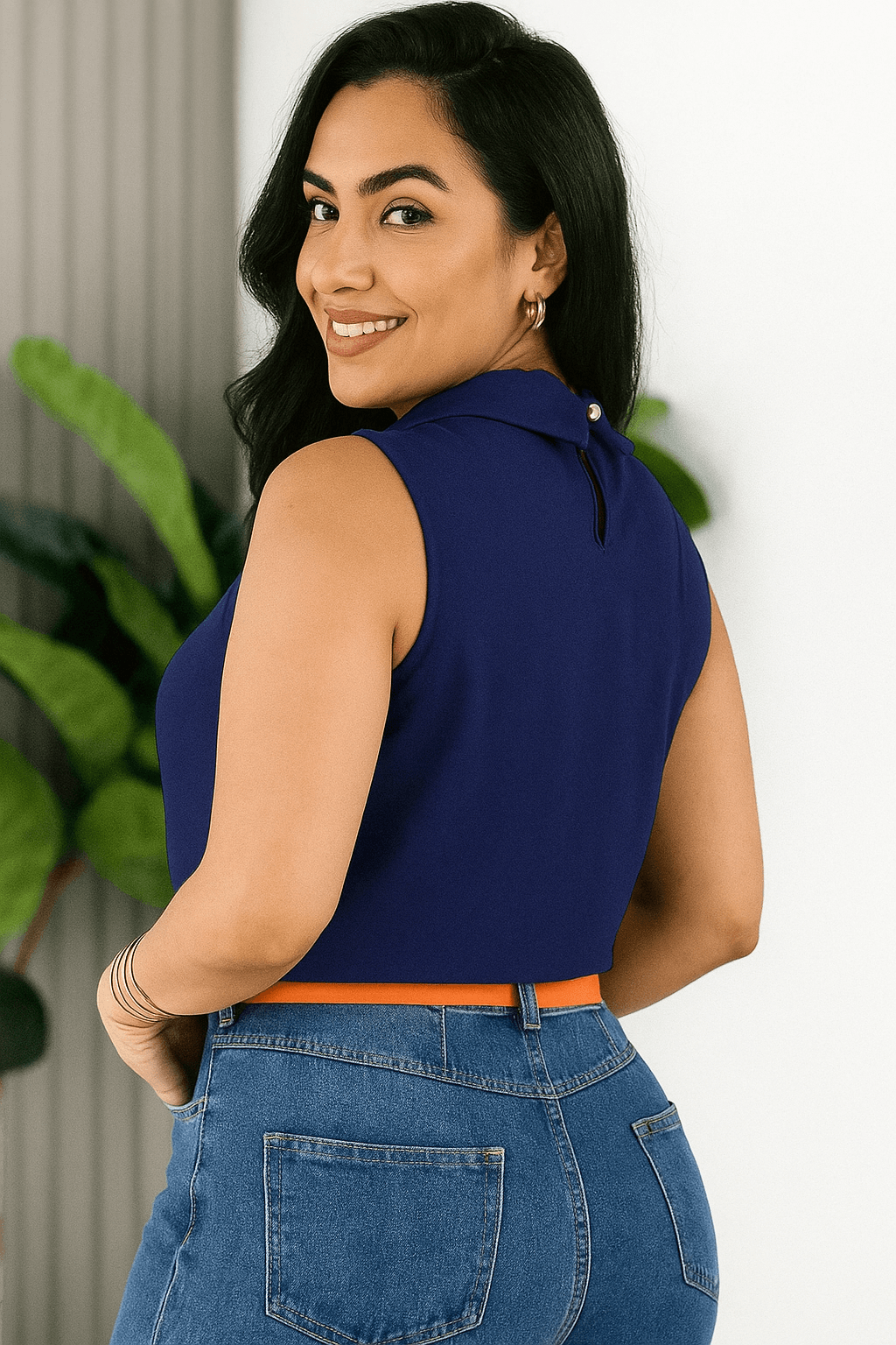 Mulher sorridente vestindo uma blusa social feminina azul marinho, sem mangas, com gola alta e detalhe plissado no busto. Ela usa calça jeans de cintura alta com cinto laranja vibrante e esta de lado. Look moderno e elegante, ideal para ambiente de trabalho ou casual