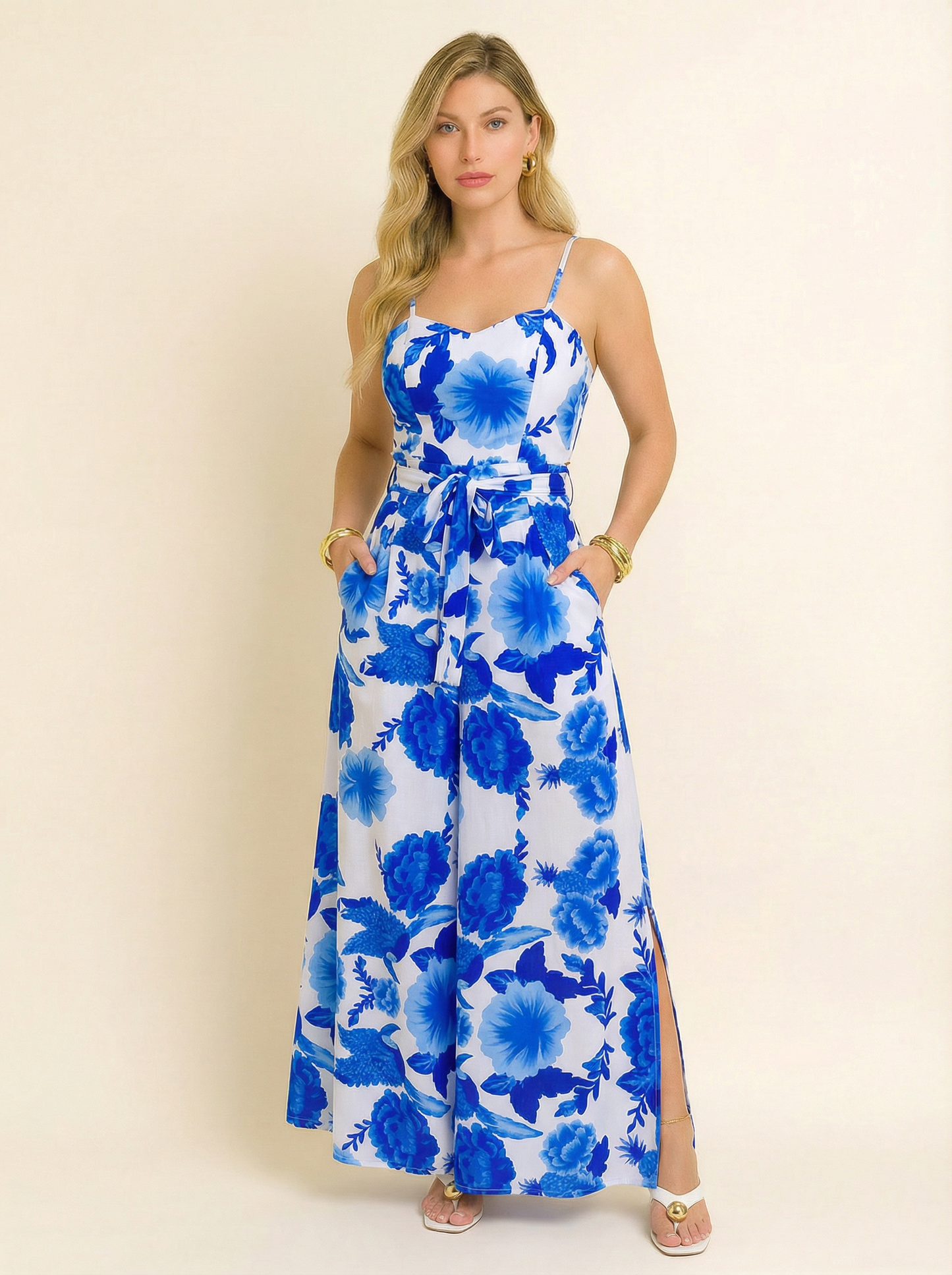 Modelo vestindo macacão longo envelope com estampa floral azul em fundo branco, com alças finas e faixa para amarração na cintura. A peça possui fenda lateral elegante e caimento fluido, ideal para ocasiões de verão com estilo e frescor.