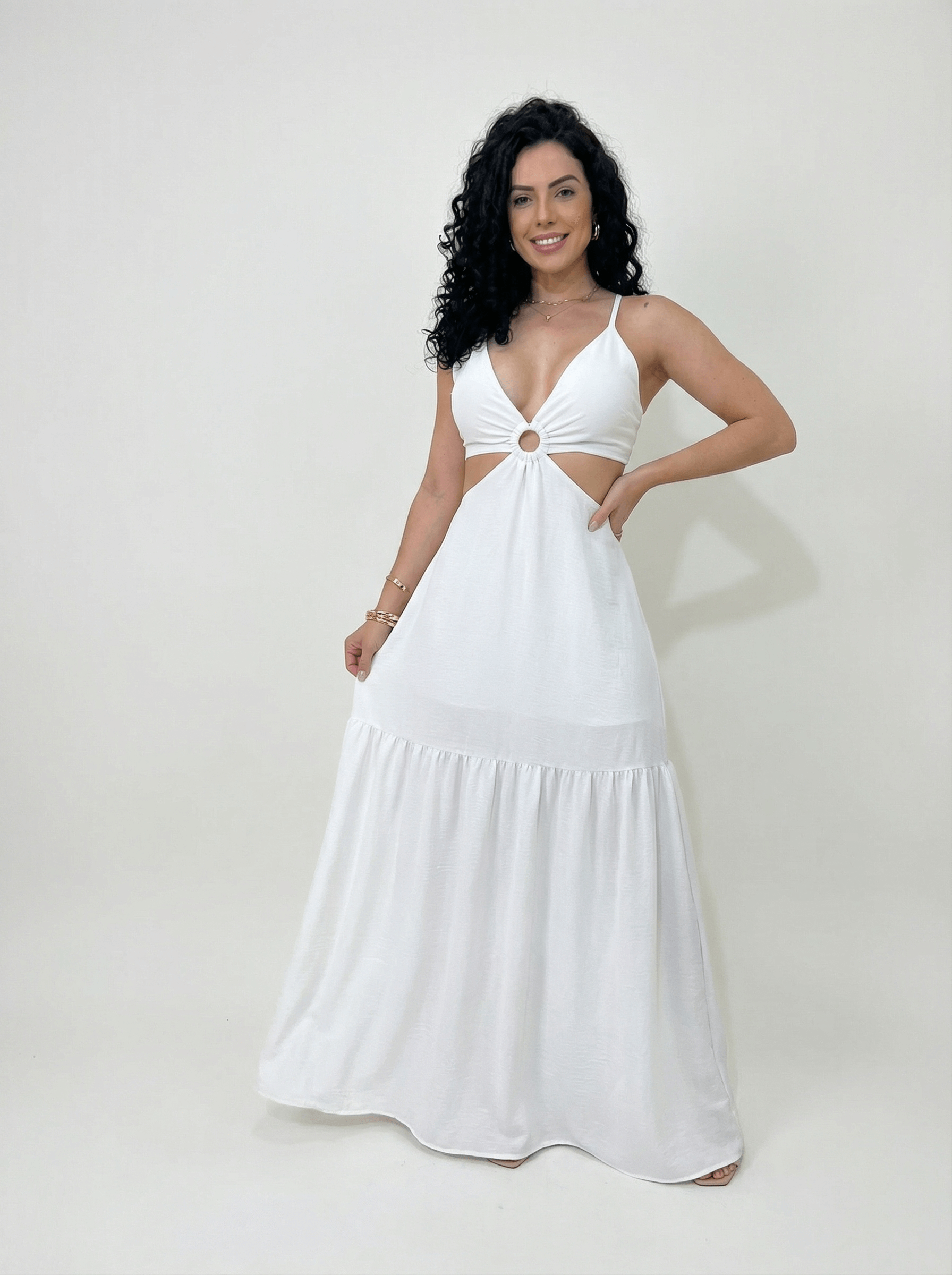 Mulher usando um vestido longo branco com decote profundo em V, alças finas e recorte lateral na cintura com detalhe em argola central. Look alegre e leve, ideal para dias quentes e ocasiões casuais de verão.