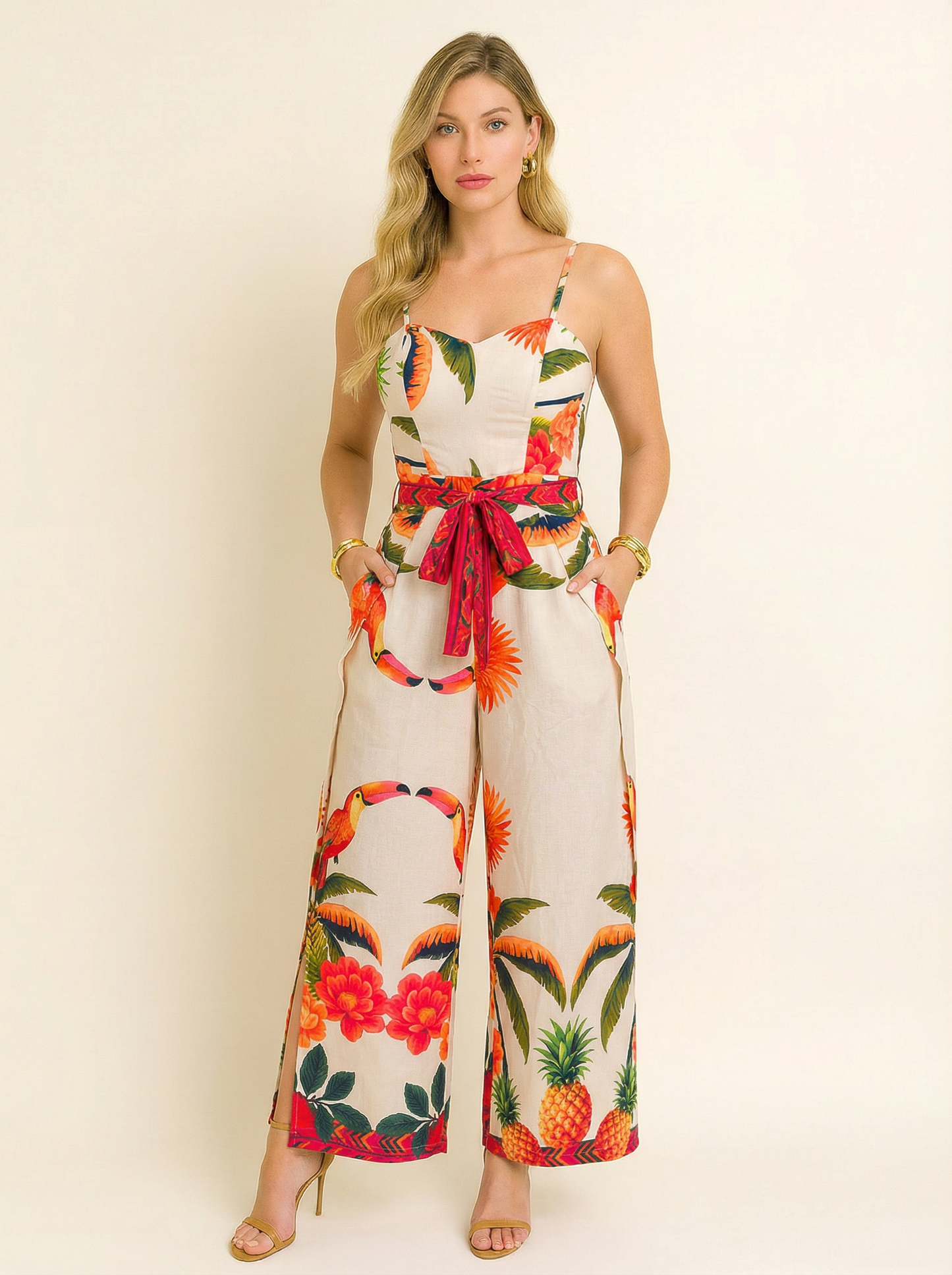 Modelo usando macacão longo com fundo bege claro e estampa tropical de abacaxis, flores e araras coloridas. A peça tem alças finas, cintura marcada com faixa vermelha estampada e caimento leve, ideal para um visual vibrante e elegante nos dias quentes de verão.