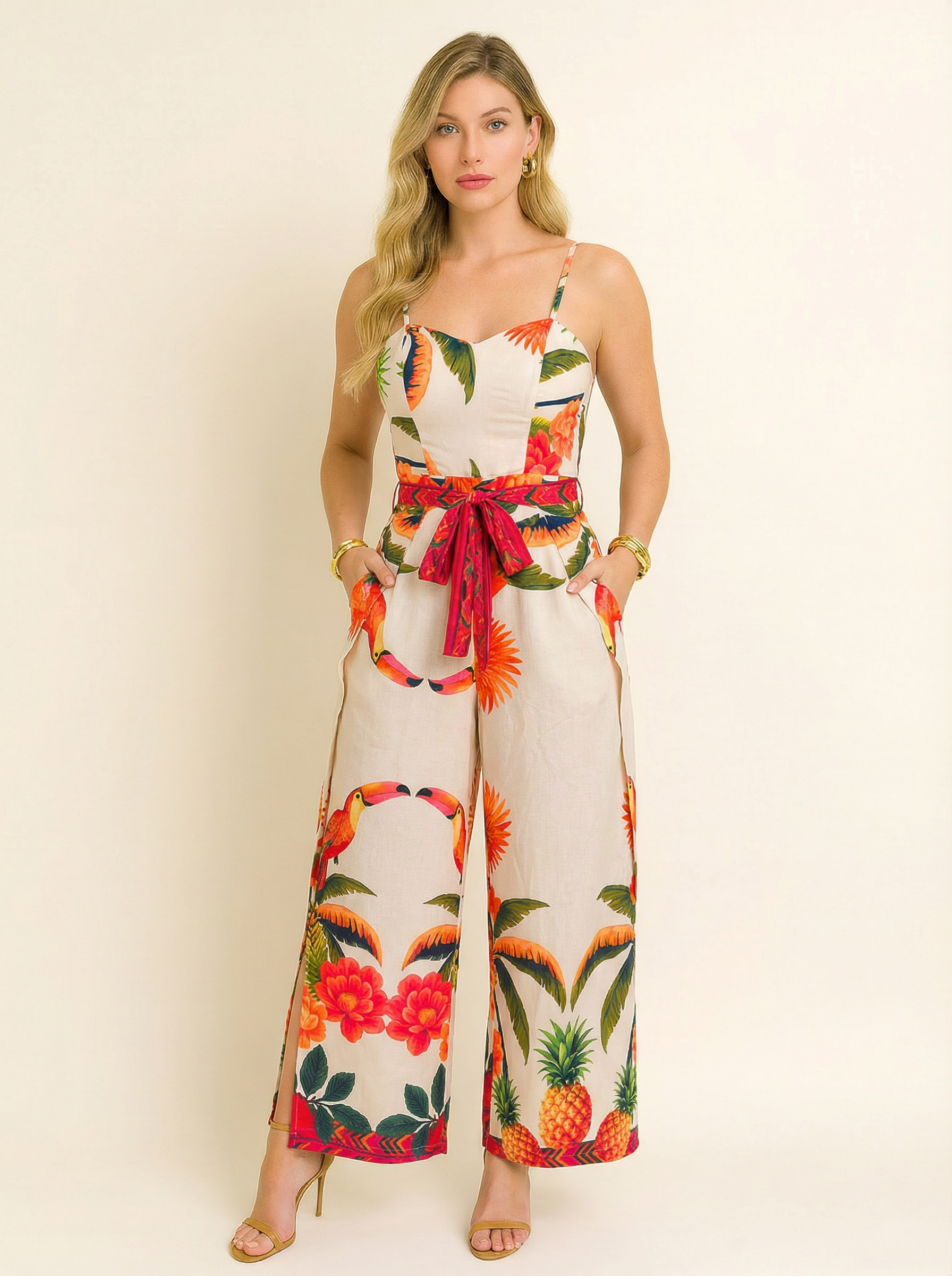Modelo usando macacão longo com fundo bege claro e estampa tropical de abacaxis, flores e araras coloridas. A peça tem alças finas, cintura marcada com faixa vermelha estampada e caimento leve, ideal para um visual vibrante e elegante nos dias quentes de verão.