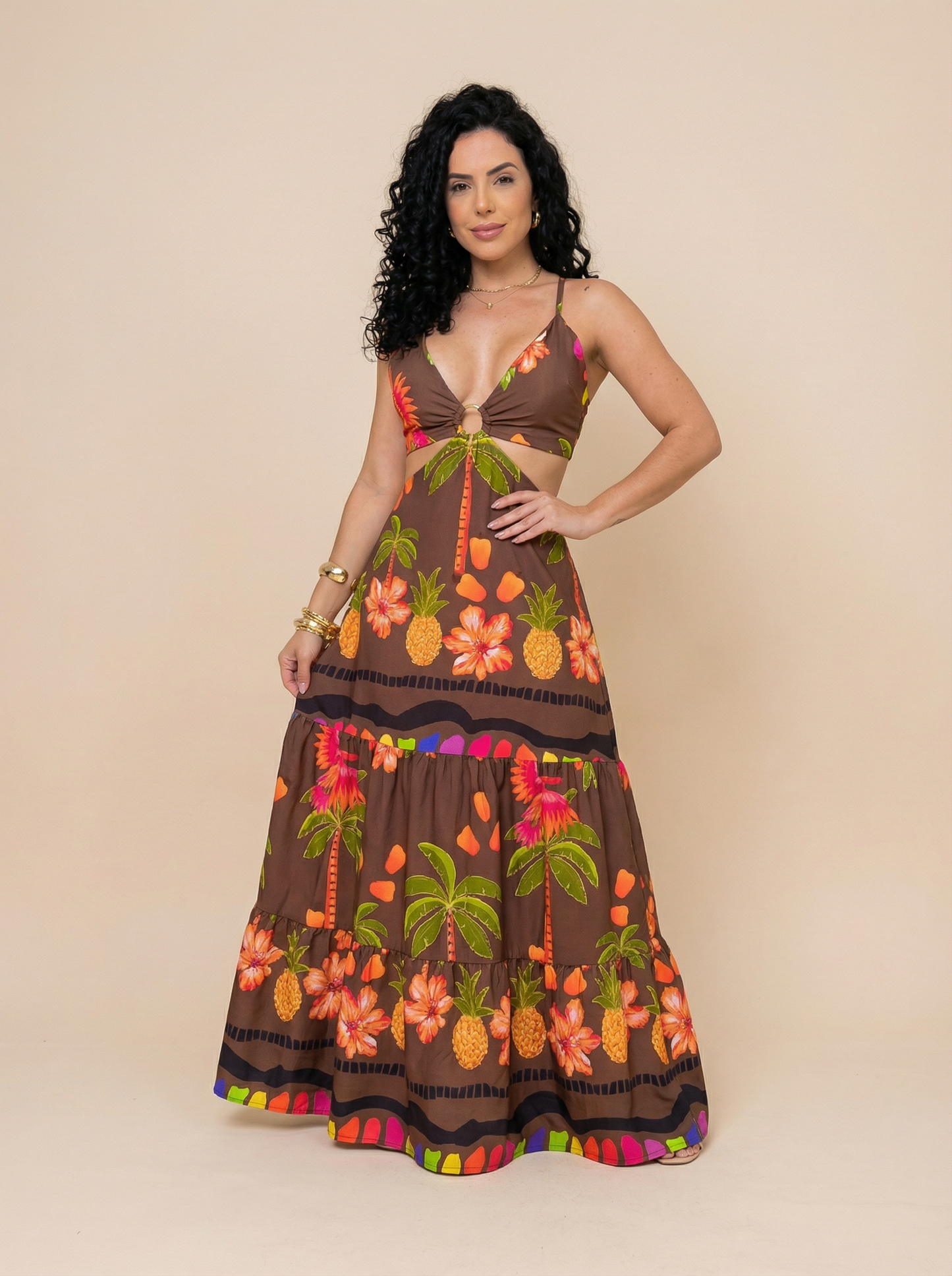 Mulher usando um vestido longo floral marrom com decote em V profundo, alças finas e abertura lateral na cintura. A estampa tropical inclui flores, coqueiros e abacaxis em tons vibrantes de laranja, verde e rosa. Ideal para o verão e ocasiões ao ar livre.