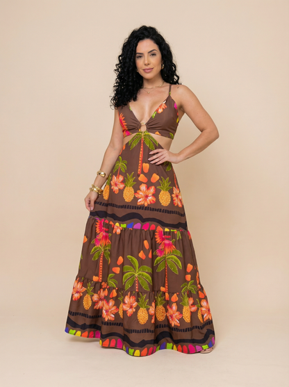Mulher usando um vestido longo floral marrom com decote em V profundo, alças finas e abertura lateral na cintura. A estampa tropical inclui flores, coqueiros e abacaxis em tons vibrantes de laranja, verde e rosa. Ideal para o verão e ocasiões ao ar livre.