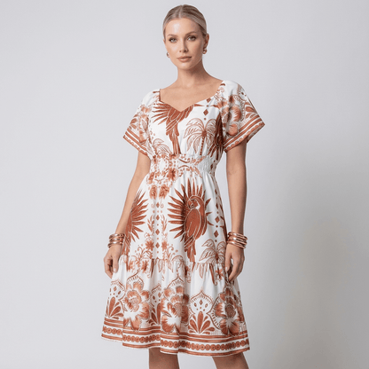 Mulher vestindo vestido midi branco com estampa marrom estilo tropical, mangas curtas bufantes, decote coração e cintura com elástico lastex. Look casual elegante ideal para dias quentes ou passeios ao ar livre.
