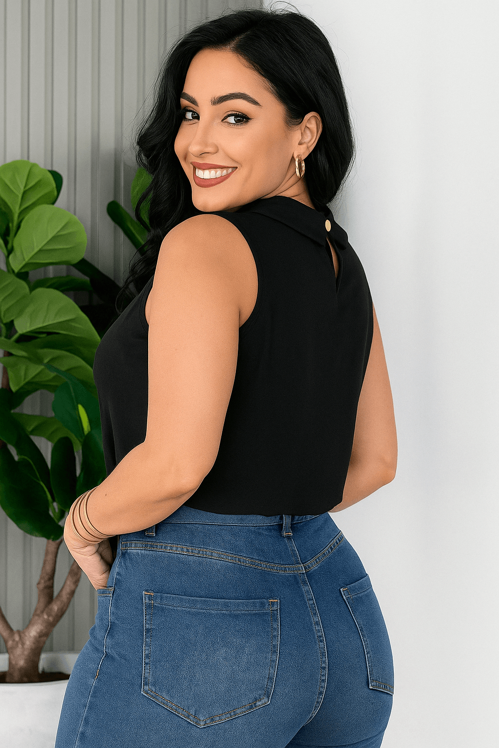 Mulher sorridente vestindo uma blusa social feminina preta, sem mangas, com gola alta e detalhe plissado no busto. Ela usa calça de alfaiataria caramelo com cinto embutido e acessórios dourados e está de lado. O ambiente é claro e elegante, com planta decorativa ao fundo