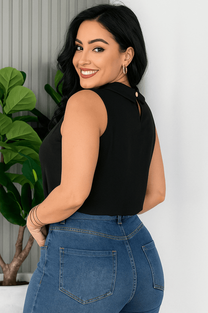 Mulher sorridente vestindo uma blusa social feminina preta, sem mangas, com gola alta e detalhe plissado no busto. Ela usa calça de alfaiataria caramelo com cinto embutido e acessórios dourados e está de lado. O ambiente é claro e elegante, com planta decorativa ao fundo