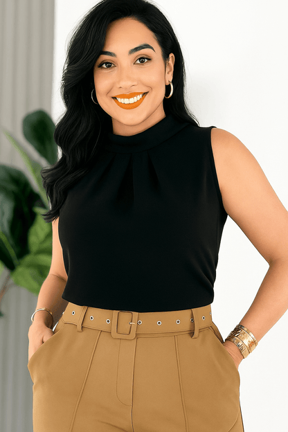 Mulher sorridente vestindo uma blusa social feminina preta, sem mangas, com gola alta e detalhe plissado no busto. Ela usa calça de alfaiataria caramelo com cinto embutido e acessórios dourados. O ambiente é claro e elegante, com planta decorativa ao fundo