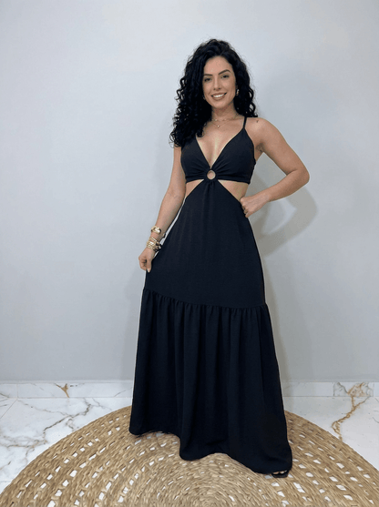 Mulher usando um vestido longo preto com decote profundo em V, alças finas e recorte lateral na cintura com detalhe em argola central. Look alegre e leve, ideal para dias quentes e ocasiões casuais de verão.