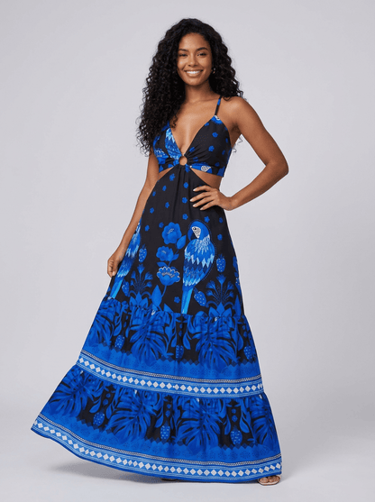 Mulher vestindo vestido longo preto com estampa tropical azul vibrante, incluindo flores e araras. Modelo com decote em V profundo, detalhe em argola no busto e recorte lateral na cintura. Look marcante e elegante para o verão.