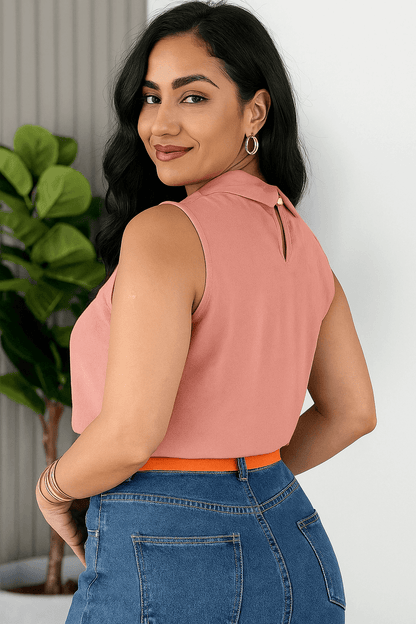 Mulher sorridente com blusa social feminina rosa bebê, sem mangas, gola alta e detalhe plissado. Look romântico e moderno, combinado com jeans azul e cinto laranja que adiciona contraste vibrante, modelo de lado.