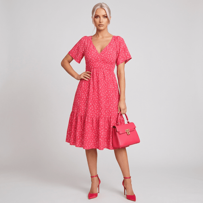 Mulher usando vestido midi rosa com estampa floral delicada, decote em V e mangas curtas. Look completo com salto alto e bolsa rosa combinando, ideal para ocasiões casuais ou encontros durante o dia – Fernanza.