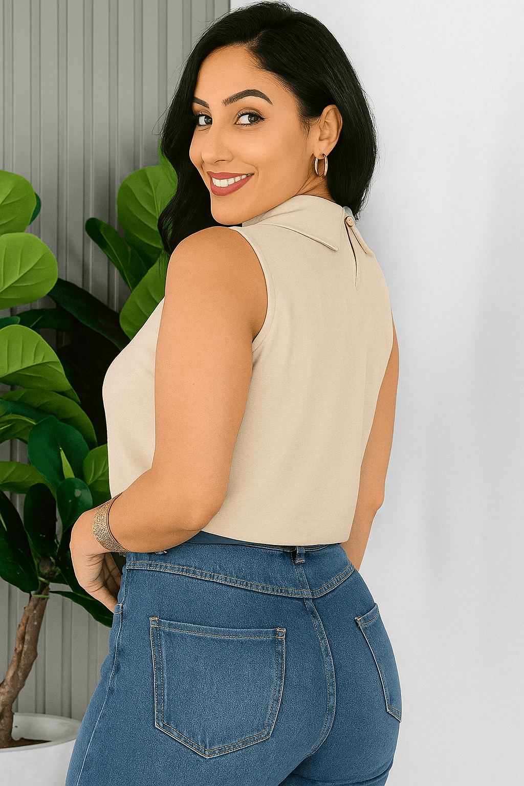 Mulher sorridente usando uma blusa social feminina bege, sem mangas, gola alta com detalhe plissado no busto. Ela combina a peça com calça jeans azul e cinto de estampa animal printe esta de lado, além de acessórios dourados. Ao fundo, um ambiente moderno com plantas verdes.